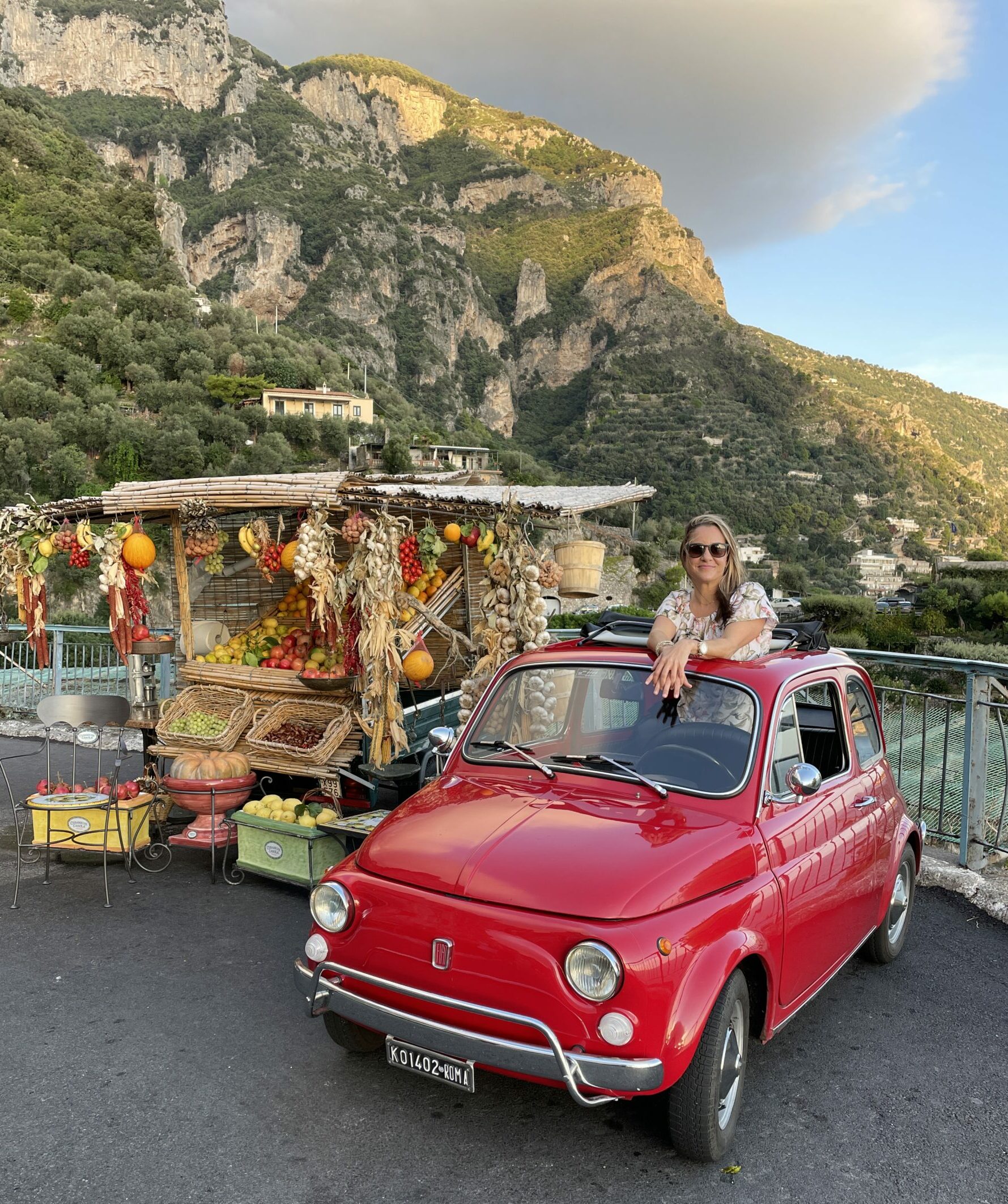 a woman is standing up through the roof of a red 1972 Fiat 500 that is parked next to a fruit and vegetable stand along the Amalfi Coast
