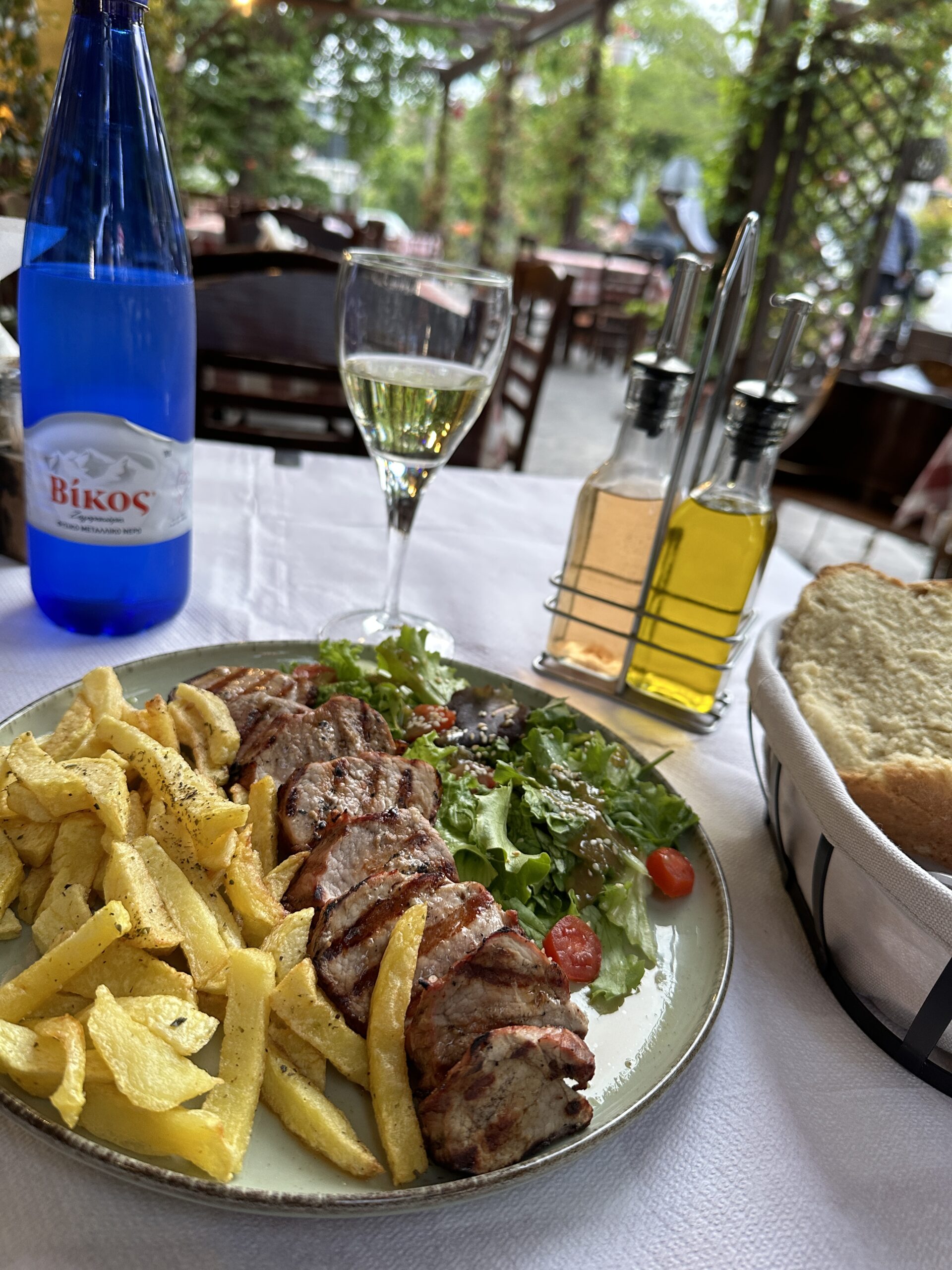 a table is shown with a plate of pork tenderloin with french fries and salad.  A basket of bread, olive oil, glass of white wine and bottle of water is shown at Yamas Taverna in Meteora Greece